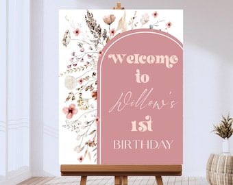 May include: A wooden easel displays a welcome sign for a first birthday. The sign features a pink arch with the text "Welcome to Willow's 1st Birthday." Floral watercolor illustrations in pink, brown, and beige decorate the sign.
