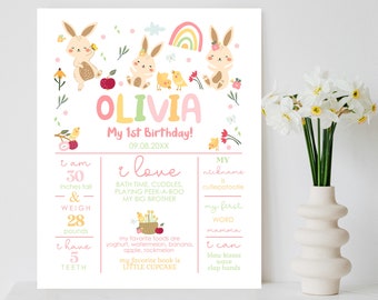 May include: A pink and green printable first birthday poster with a bunny theme. The poster features a girl's name, Olivia, and details about her first year, including her height, weight, favorite foods, and activities. The poster is decorated with bunnies, flowers, and a rainbow.