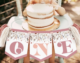 May include: A banner with the word "ONE" in large letters, with a floral design, hangs in front of a baby eating a cake. The banner is pink and white, with tassels on each end. The cake is white with a gold cake stand.