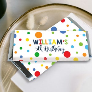 May include: A candy bar wrapper with a white background and colorful polka dots. The text "WILLIAM'S 5th Birthday" is displayed in a playful font. The wrapper is partially unwrapped, revealing the silver foil inside.