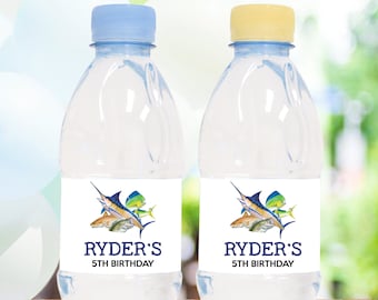 May include: Two clear water bottles with blue and yellow caps. Each bottle has a white label with a watercolor illustration of fish and the text "Ryder's 5th Birthday" in navy blue. The background is blurred.