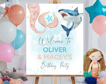 Op de afbeelding: Een welkomstbord voor een verjaardagsfeestje op een ezel, met een zeemeermin- en haaienthema. Op het bord staat "Welcome to Oliver & Macey's Birthday Party". Ballonnen in roze, blauw en wit zijn zichtbaar, samen met een zeemeerminnenpop.