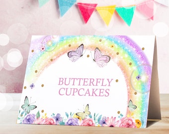 Peut inclure: Une pancarte blanche avec un arc-en-ciel et des papillons, avec le texte "BUTTERFLY CUPCAKES" en lettres roses. La pancarte est décorée de paillettes dorées et de fleurs.