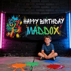 Könnte beinhalten: Ein schwarzes Banner mit den Worten "HAPPY BIRTHDAY MADDOX" in weißer und farbenfroher Schrift. Die Zahl "67" wird mit farbenfrohen Farbspritzern dargestellt. Eine junge Person sitzt vor dem Banner.