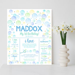 Puede incluir: Un cartel de primer cumpleaños con temática de burbujas azules y amarillas para un niño llamado Maddox. El cartel incluye detalles como su edad, altura, peso, comidas favoritas y primeras palabras.