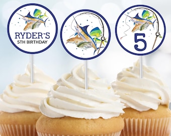 Puede incluir: Tres cupcakes cubiertos con glaseado blanco y adornos para pasteles. Los adornos presentan un diseño circular azul y blanco con una ilustración de acuarela de peces y aparejos de pesca. Un adorno dice "Ryder's 5th Birthday", y otro muestra el número "5".