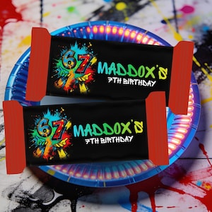 Könnte beinhalten: Zwei schwarze Schokoriegelverpackungen mit roten Enden werden auf einem blauen Teller präsentiert. Die Verpackungen zeigen einen farbenfrohen Text im Graffiti-Stil mit der Aufschrift "67 MADDOX'S 7TH BIRTHDAY". Der Teller ist mit farbenfrohen Farbspritzern verziert.