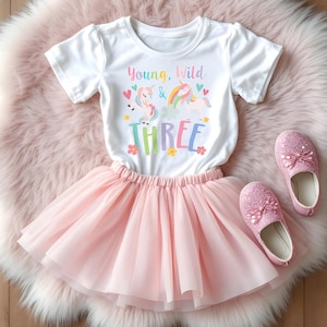 May include: A white t-shirt with the text "Young, Wild & THREE" and unicorn graphics, paired with a pink tutu skirt and glittery pink shoes. The outfit is designed for a child's third birthday.