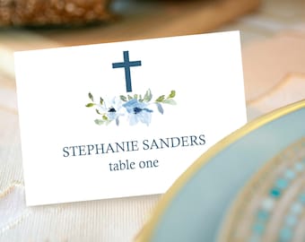 May include: White place card with a blue cross and floral design. The text reads "Stephanie Sanders table one."