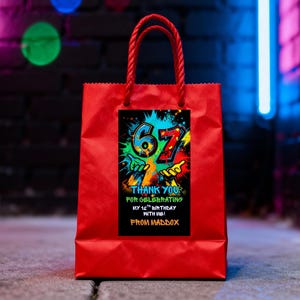 May include: A red gift bag with a twisted red rope handle. The bag features a black rectangular label with colorful graffiti-style text that reads "67" and "Thank You for Celebrating My 12th Birthday with Me! From Maddox."