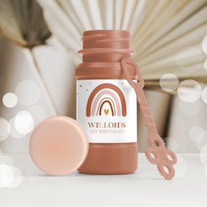 May include: A brown plastic bottle of bubble solution with a pink lid and a pink wand with a bubble ring. The bottle has a white label with a rainbow design and the text "WILLOH'S 1ST BIRTHDAY".