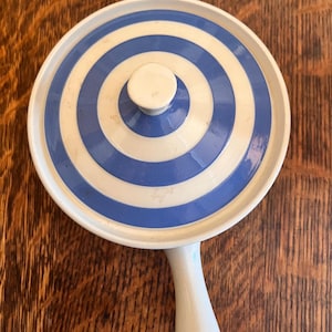 May include: A white ceramic serving dish with a blue and white striped lid and a white handle. The lid features concentric circles of blue and white. The dish is set on a wooden surface.