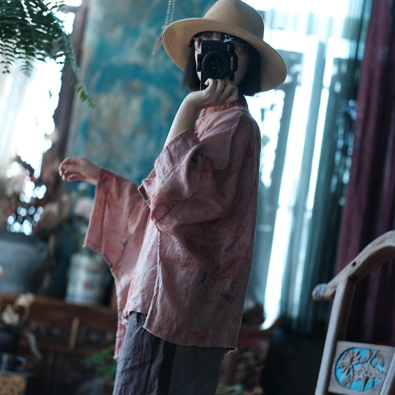 May include: A light pink, loose-fitting blouse with wide sleeves and a subtle mottled pattern, paired with dark grey trousers and a tan wide-brimmed hat. The garment features a high neckline and a relaxed silhouette, captured in a side profile view.