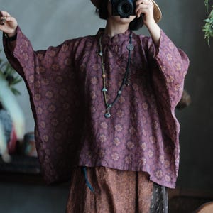May include: A loose-fitting, plum-colored blouse featuring a repeating floral pattern in muted yellow and brown. The garment has wide, three-quarter length sleeves, a high mandarin-style collar with a side button closure, and a straight hemline.