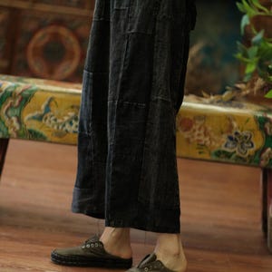 May include: Wide-leg cropped trousers in a dark charcoal gray denim fabric with visible vertical panel stitching and a relaxed fit, paired with dark olive green slip-on shoes featuring a rounded toe and visible contrast stitching along the sole.