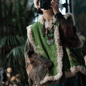May include: A green vest featuring a subtle floral pattern and thick, cream-colored faux fur trim along the edges. The garment includes dark brown, textured patchwork sleeves and is worn with a layered necklace featuring a large, pale green pendant.