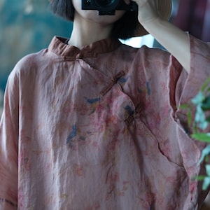 May include: A dusty rose-coloured blouse featuring a mandarin collar and an asymmetrical front closure secured with fabric toggle buttons. The garment is decorated with a delicate print of small blue birds and soft pink floral branches across the fabric.