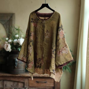 May include: An olive green loose-fitting top featuring a faded floral print with pink roses and dark green leaves. The garment has a round neckline, wide three-quarter length sleeves, and a raw, uneven hemline with visible dark brown inner lining.