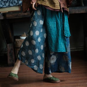 May include: Wide-leg trousers with a distinctive design. One leg has large white polka dots on a navy blue background, the other a teal panel and floral print. The trousers are worn with green patterned shoes.