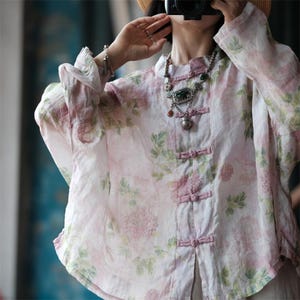 Puede incluir: Una blusa de lino rosa claro con un estampado floral en tonos rosas y verdes. La blusa presenta un cuello mandarín, mangas largas y cierres de nudo. Se ve un collar plateado con un colgante.