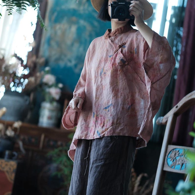 May include: A dusty rose-coloured loose-fitting blouse with a mandarin collar, side button closure, and long sleeves. The fabric features a subtle, abstract watercolour-style print in muted blue and pink tones, paired with dark brown wide-leg trousers.