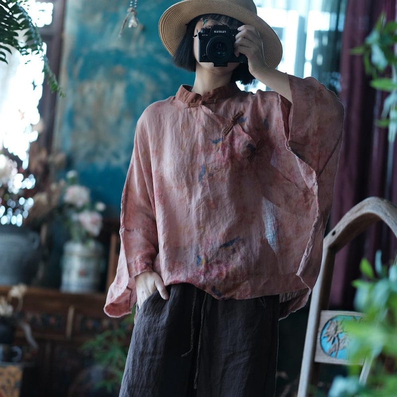May include: A loose-fitting, dusty rose-coloured blouse with a mandarin collar and subtle, multi-coloured paint-like splatters. The garment features wide, three-quarter length sleeves and is paired with dark brown, drawstring-waist trousers.