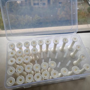 以下が含まれることがあります： 透明なプラスチック製の収納ボックスに、多数の白い糸巻きが入っています。糸巻きはボックス内にきちんと配置されており、透明な蓋が付いています。糸巻きは円筒形で、円形の上面と底面があります。