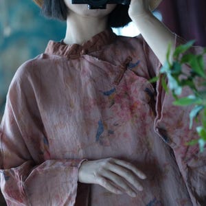 May include: A dusty rose-coloured long-sleeve top featuring a mandarin collar with a toggle closure. The fabric is adorned with a subtle, repeating print of small blue birds perched on branches amidst soft pink floral blossoms across the garment.