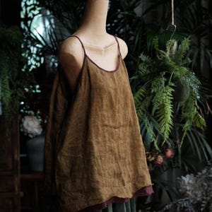 May include: A sleeveless tank top in a muted olive-brown color, featuring thin spaghetti straps and a scoop neckline. The garment has a relaxed, loose-fitting silhouette with a visible dark maroon or deep red trim along the neckline and hem.