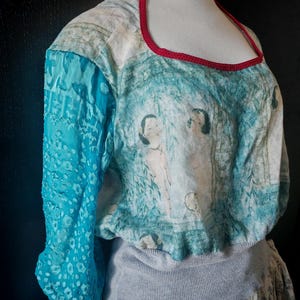 May include: A unique top with a square neckline and a red trim. The top features a blue and white watercolor-style print with figures and a turquoise sleeve with a floral pattern. The bottom portion is a light gray sweater material.