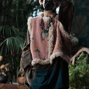 Puede incluir: Un atuendo en capas con un chaleco rosa con ribete de piel sintética color crema. El chaleco, con estampado floral, se lleva sobre una parte superior estampada y una falda verde oscuro. Una cámara se sostiene frente a la cara.