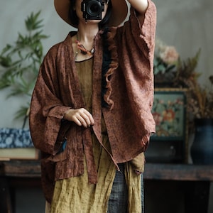 May include: A brown patterned open-front jacket with wide sleeves and a black ruffled trim along the collar. The jacket is worn over a mustard yellow textured top. A wide-brimmed straw hat is visible at the top of the frame.