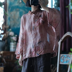 May include: A loose-fitting, long-sleeved blouse in a muted dusty pink hue with a subtle, mottled watercolour-style pattern in shades of blue and orange. The garment features a mandarin collar, a side-fastening placket with fabric toggles, and wide sleeves.