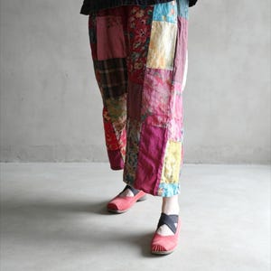 Puede incluir: Pantalones anchos con diseño de patchwork que presenta varios cuadrados de tela floral, a cuadros y lisos en tonos rojos, rosas, azules y amarillos. Los pantalones se combinan con zapatos rojos sin cordones con tiras elásticas negras cruzadas.
