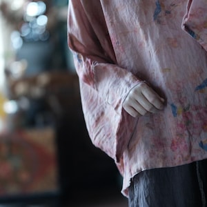 May include: A loose-fitting, light pink top with long, wide sleeves and a subtle, multicoloured floral and bird print. The fabric has a textured, slightly wrinkled appearance, and the garment is paired with dark grey trousers.