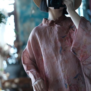 May include: A dusty rose long-sleeved blouse featuring a mandarin collar with a small fabric toggle closure. The garment is decorated with a subtle, muted floral print in shades of blue and soft pink, crafted from a textured, lightweight fabric.
