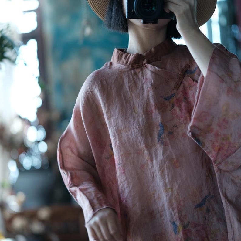 May include: A dusty rose long-sleeved blouse featuring a mandarin collar with a small fabric toggle closure. The garment is decorated with a subtle, muted floral print in shades of blue and soft pink, crafted from a textured, lightweight fabric.