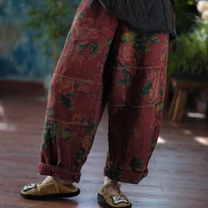 May include: Loose-fitting trousers in a muted brick red colour featuring a prominent floral pattern with green leaves and pink blossoms. The trousers have a visible horizontal seam across the mid-thigh area and cuffed hems at the bottom of the legs.