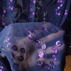 Puede incluir: Una tela azul marino transparente con un patrón repetitivo de pequeños lunares metálicos de color púrpura. El material ligero es translúcido, permitiendo el paso de la luz y resaltando la textura de la tela.