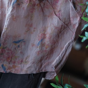 May include: A light pink, loose-fitting top featuring a delicate print of small blue birds perched among clusters of soft pink and yellow flowers. The fabric has a visible, slightly textured weave and a relaxed, draped silhouette with a hemline.