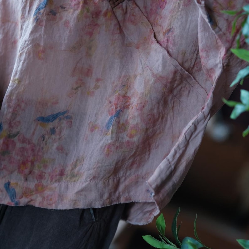 May include: A light pink, loose-fitting top featuring a delicate print of small blue birds perched among clusters of soft pink and yellow flowers. The fabric has a visible, slightly textured weave and a relaxed, draped silhouette with a hemline.