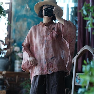 May include: A loose-fitting, dusty pink blouse with a mandarin collar and wide, three-quarter length sleeves. The fabric features a subtle, mottled pattern with faint blue and yellow accents. It is paired with dark brown, wide-leg trousers.