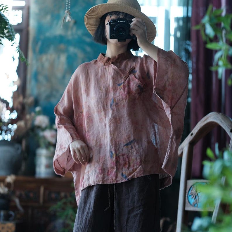 May include: A loose-fitting, dusty pink blouse with a mandarin collar and wide, three-quarter length sleeves. The fabric features a subtle, mottled pattern with faint blue and yellow accents. It is paired with dark brown, wide-leg trousers.