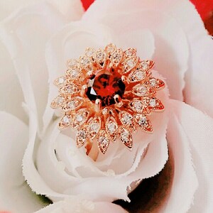 以下が含まれることがあります： 赤い宝石が、小さな透明な宝石で囲まれた金の指輪。指輪は、たくさんの花びらを持つ花の形をしています。