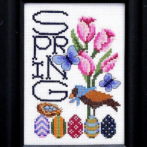 Könnte beinhalten: Kreuzstich-Stickerei mit einer Frühlingsszene mit dem Wort "FRÜHLING" in vertikalen Buchstaben, rosa Tulpen, blauen Schmetterlingen, einem braunen Vogel mit einem blauen Band und einem Nest mit blauen Eiern. Unter dem Vogel befinden sich fünf bunte Ostereier.