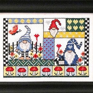 Könnte beinhalten: Eine Kreuzstich-Stickerei mit drei Gnomen, die blaue und weiße Hüte und Bärte tragen. Die Gnome stehen vor einem bunten Patchwork-Hintergrund mit roten Blumen und einem Schmetterling.