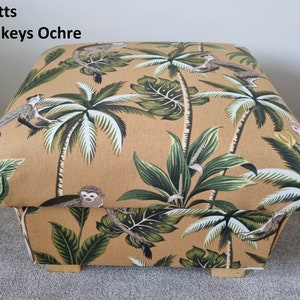 Puede incluir: Un puf de almacenamiento de tela beige con un estampado de mono y palmeras. La tela tiene una textura sutil y el puf tiene patas de madera.