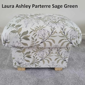 Könnte beinhalten: Ein quadratischer Hocker, bezogen mit Stoff "Laura Ashley Parterre Sage Green". Der Stoff hat ein Blumenmuster mit weißen, salbeigrünen und grauen Blumen und Blättern. Der Hocker steht auf vier kleinen, hellen Holzfüßen.