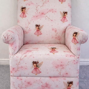 May include: Pink upholstered armchair with a fairy and floral pattern. The chair features a light pink background with illustrations of fairies in pink dresses and floral accents. The chair has wooden legs.