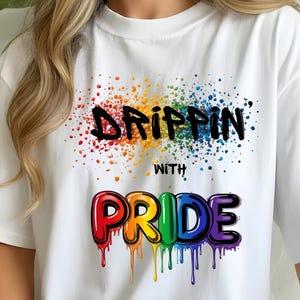 Op de afbeelding: Wit T-shirt met de tekst "DRIPPIN' WITH PRIDE" in een kleurrijk, druipend lettertype. Het ontwerp heeft een regenboogkleurig spatteffect boven de tekst, waardoor een levendige afbeelding ontstaat.